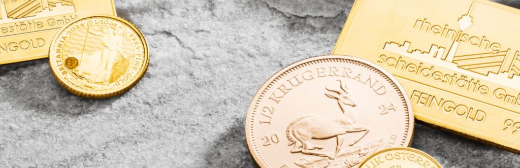 Goldbarren und Goldmünzen, darunter Krügerrand und Britannia, auf grauer Steinoberfläche. Die Aufnahme steht für den Kauf von Anlagegold und Edelmetallen bei der Rheinischen Scheidestätte.