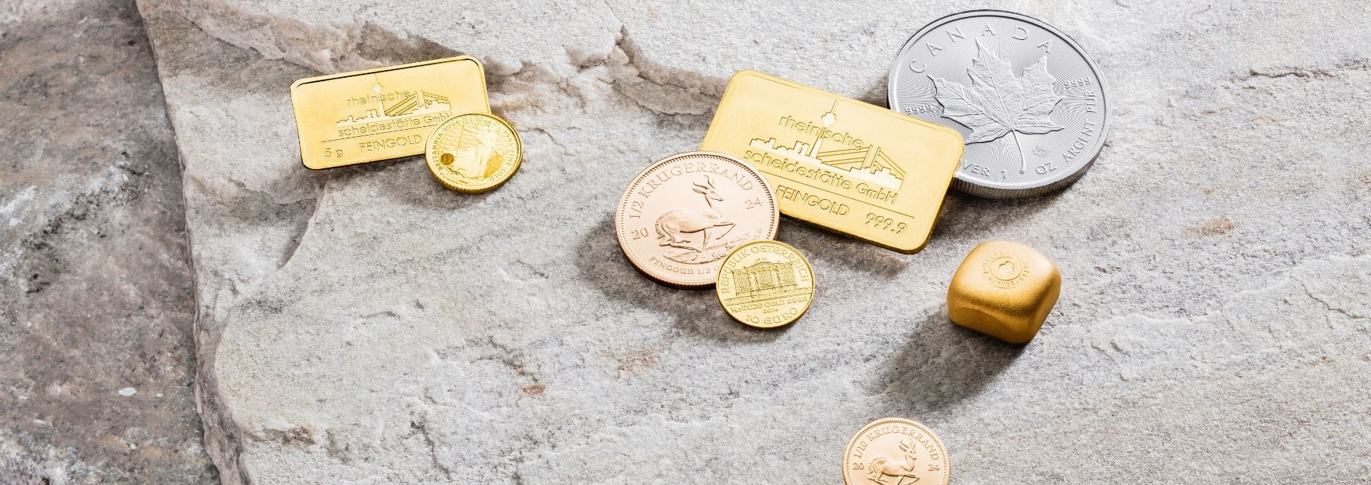 Feingoldbarren, Anlagemünzen und Silbermünzen in unterschiedlicher Größe auf Naturstein. Die Darstellung veranschaulicht den Kauf von Edelmetallen wie Gold und Silber sowie die Investition in physische Sachwerte.