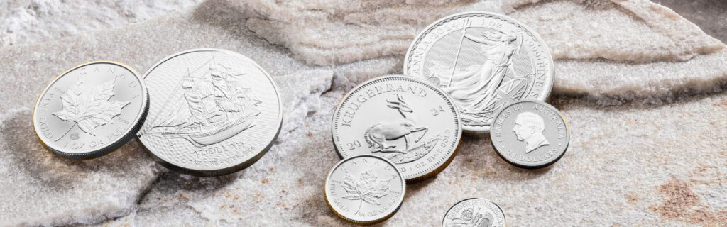 Verschiedene Anlagesilbermünzen internationaler Prägestätten auf einer Steinoberfläche, darunter Motive des Krügerrand, Britannia und Maple Leaf. Die Aufnahme symbolisiert den Kauf von Silbermünzen in Kassel und die Investition in physisches Silber als wertbeständige Edelmetallanlage.
